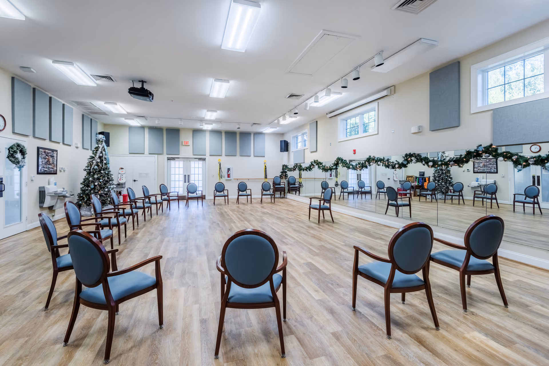 A large, bright room with wooden flooring and a circle of blue cushioned chairs with wooden frames. The room has large mirrors on one wall decorated with garlands and Christmas lights. There is a decorated Christmas tree and a snowman figure near the back wall, along with a water fountain and a clock. The ceiling has multiple fluorescent lights and a projector. Windows near the ceiling let in natural light.