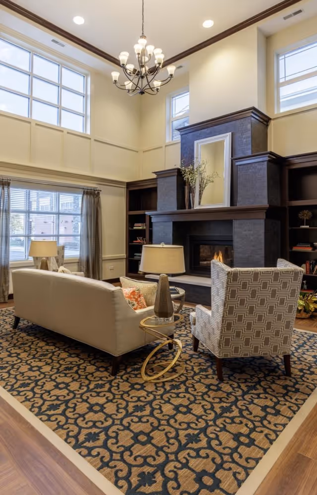 A cozy living room with a patterned area rug, a beige sofa, a patterned armchair, and a round side table with a lamp. The room features a large dark stone fireplace with a mirror and decorative vases on the mantel, built-in bookshelves on either side, tall windows letting in natural light, and a chandelier hanging from the high ceiling.