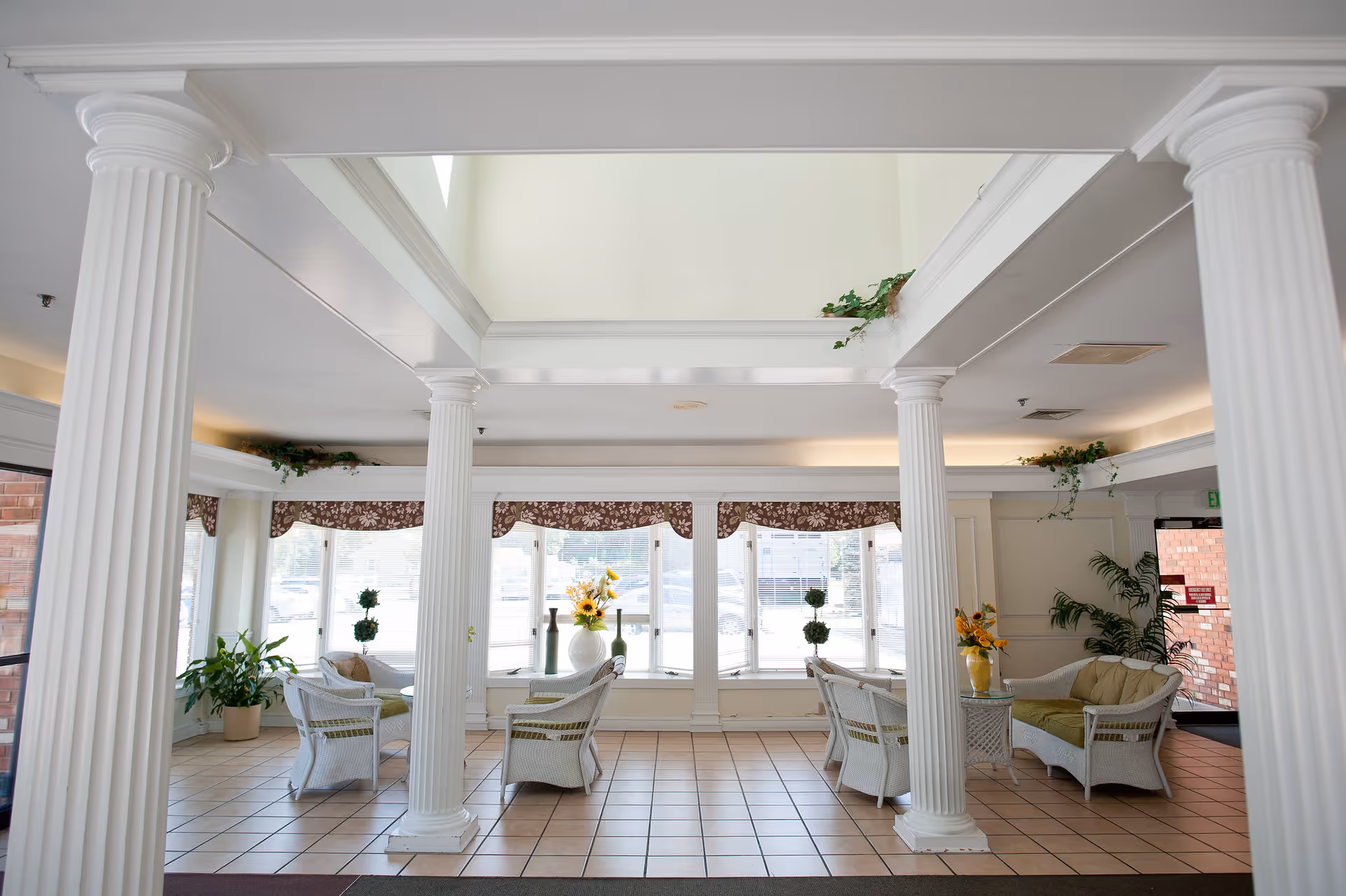 Bright and spacious seating area with white wicker chairs and sofas arranged around small tables with flower vases. The room features large windows with floral valances, white columns, tiled floor, and some green plants placed around the space.
