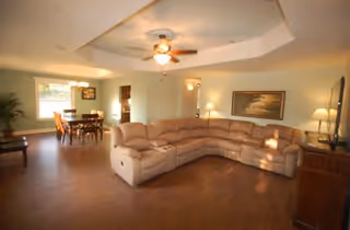 A spacious living room with a large beige sectional sofa arranged in an L-shape, a ceiling fan with lights, wooden flooring, and a dining area with a table and chairs visible in the background. The room is softly lit with natural light coming through windows and several lamps.