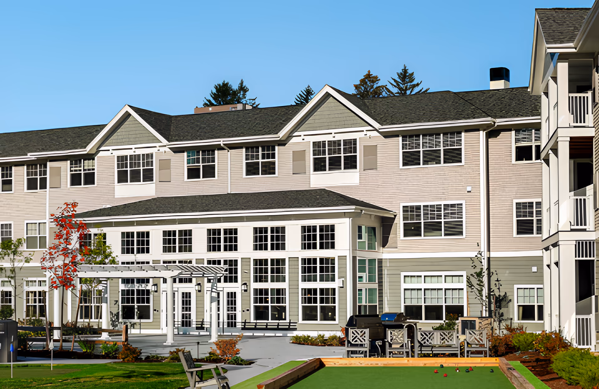 Exterior view of a multi-story retirement community building with large windows and a covered patio area. In the foreground, there is a bocce ball court with chairs and tables around it, surrounded by landscaped greenery and a clear blue sky.