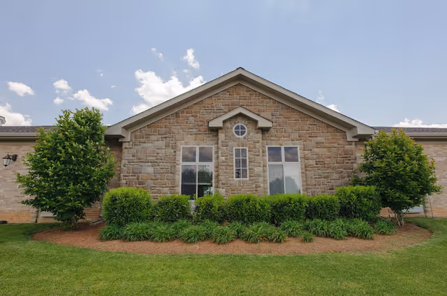 Exterior view of a single-story building with a stone facade, two large windows, and a small circular window above. The building is surrounded by neatly trimmed bushes, two small trees, and a well-maintained lawn under a partly cloudy sky.