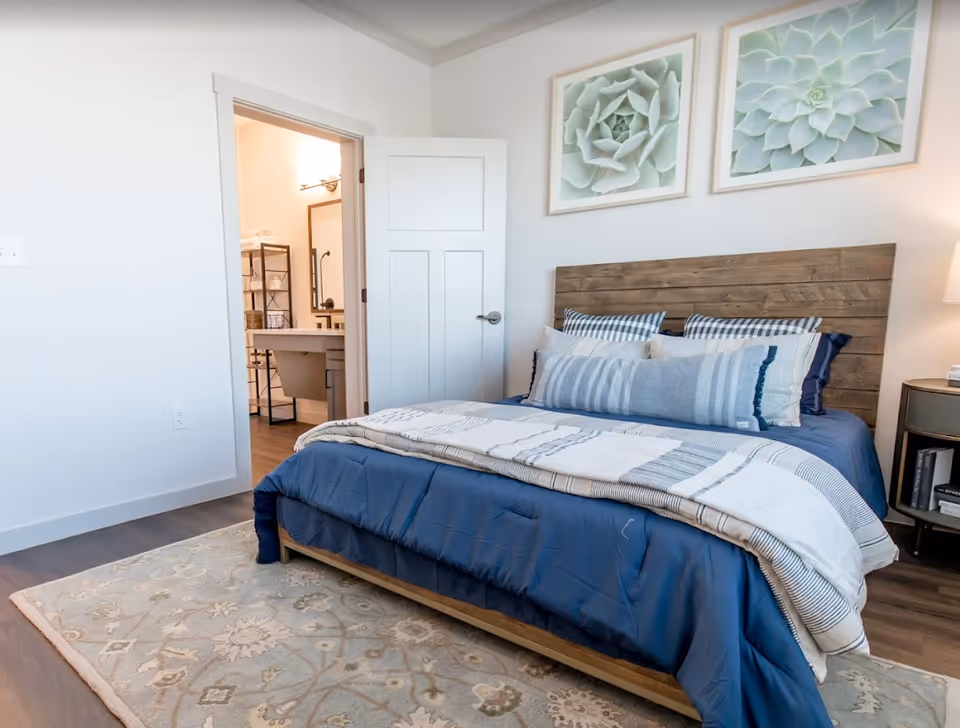 Bright bedroom with a wooden headboard and blue bedding, a rug, nightstand, and an open door leading to a bathroom.