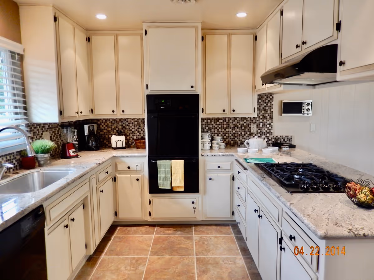 A clean and well-organized kitchen with white cabinets, a double oven, a gas stove, a sink under a window with blinds, and various kitchen appliances and utensils on the countertops. The backsplash features a mosaic tile design, and the floor is tiled in a warm brown color.