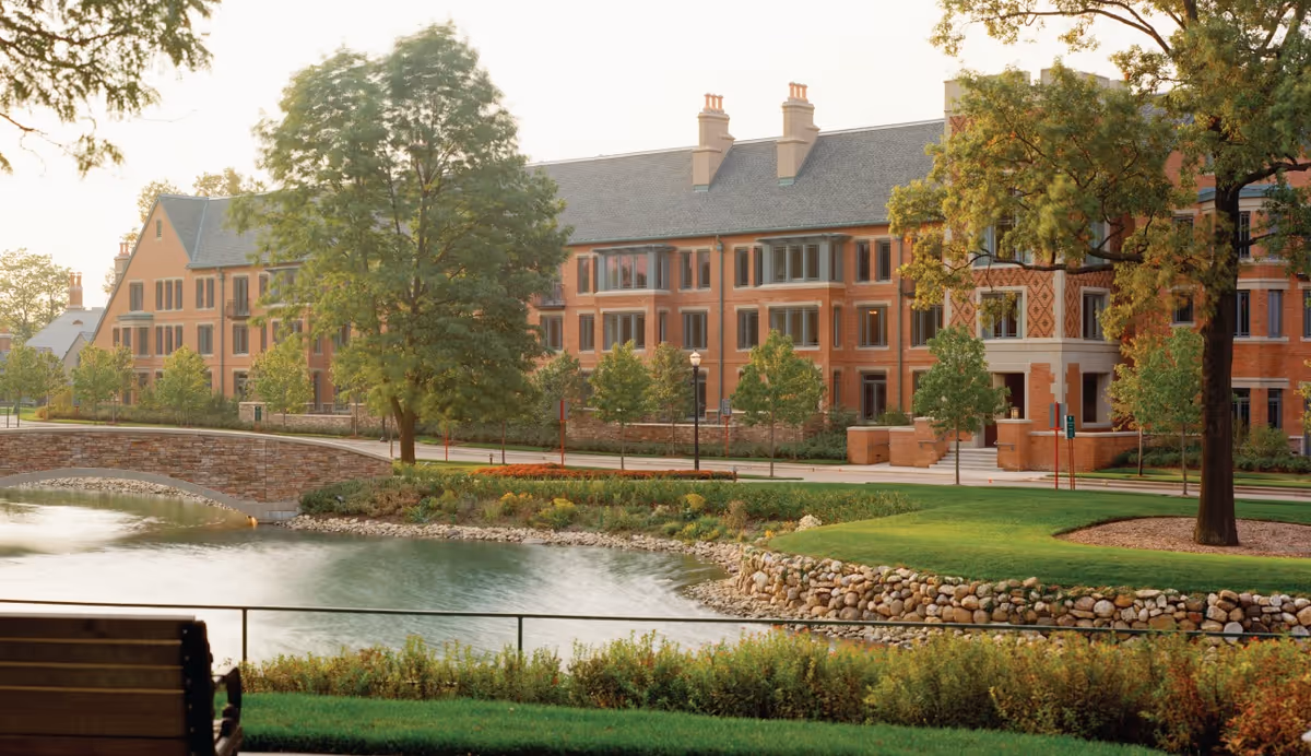 A large brick building with multiple windows and chimneys, surrounded by green trees and landscaping. In the foreground, there is a pond with a stone bridge crossing over it, and a wooden bench facing the water. The scene is peaceful and well-maintained, suggesting a senior living facility environment.