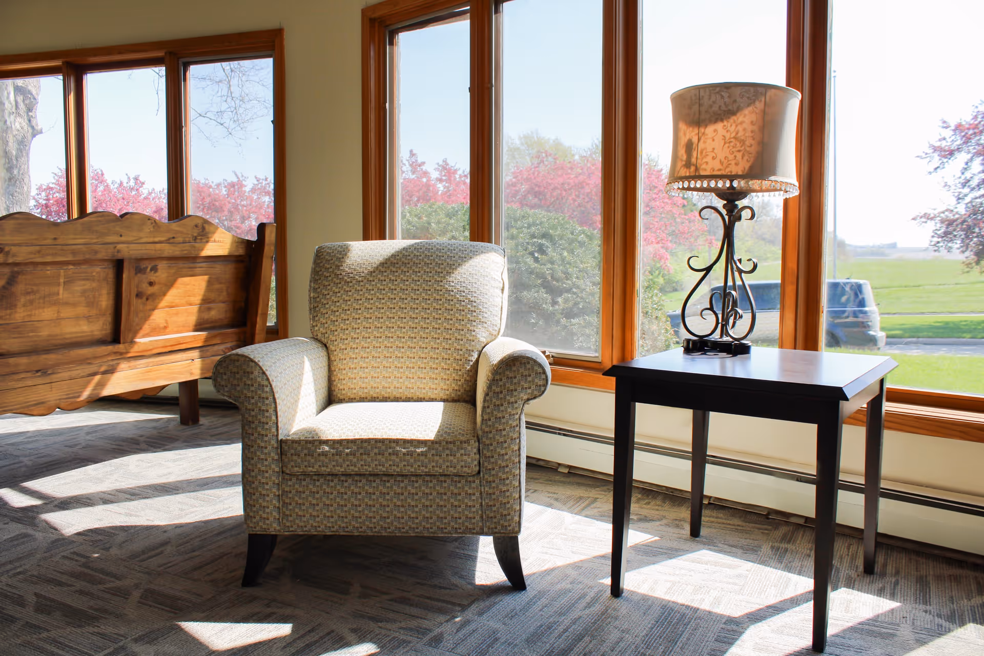 Sunlit sitting area with an upholstered armchair next to a side table and decorative lamp in front of large windows showing trees outside.