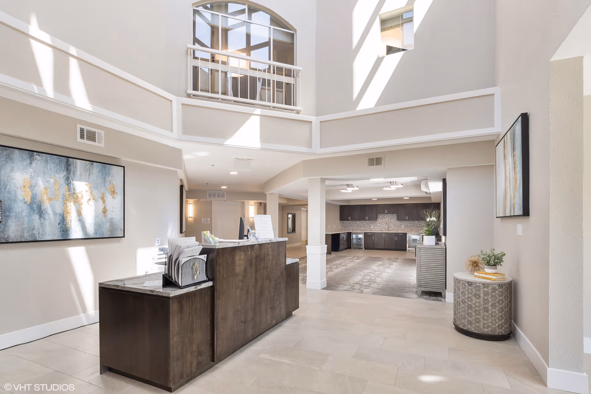 Bright and spacious interior of a senior living facility with a modern reception desk in the foreground, light-colored walls, and large windows allowing natural light to fill the space. There is a kitchen area with dark cabinets and stainless steel appliances in the background, along with decorative artwork on the walls and a small round table with plants and books on the right.