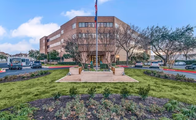 Brick senior living facility exterior with flagpole, entrance stairs, and landscaped grounds.