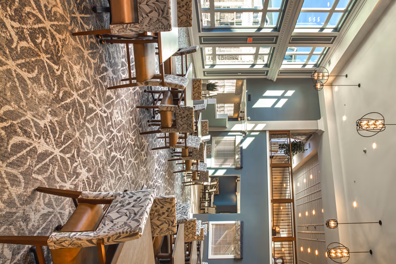 Bright, spacious dining and lounge area with patterned carpet, multiple tables and upholstered chairs, large windows and hanging pendant lights.