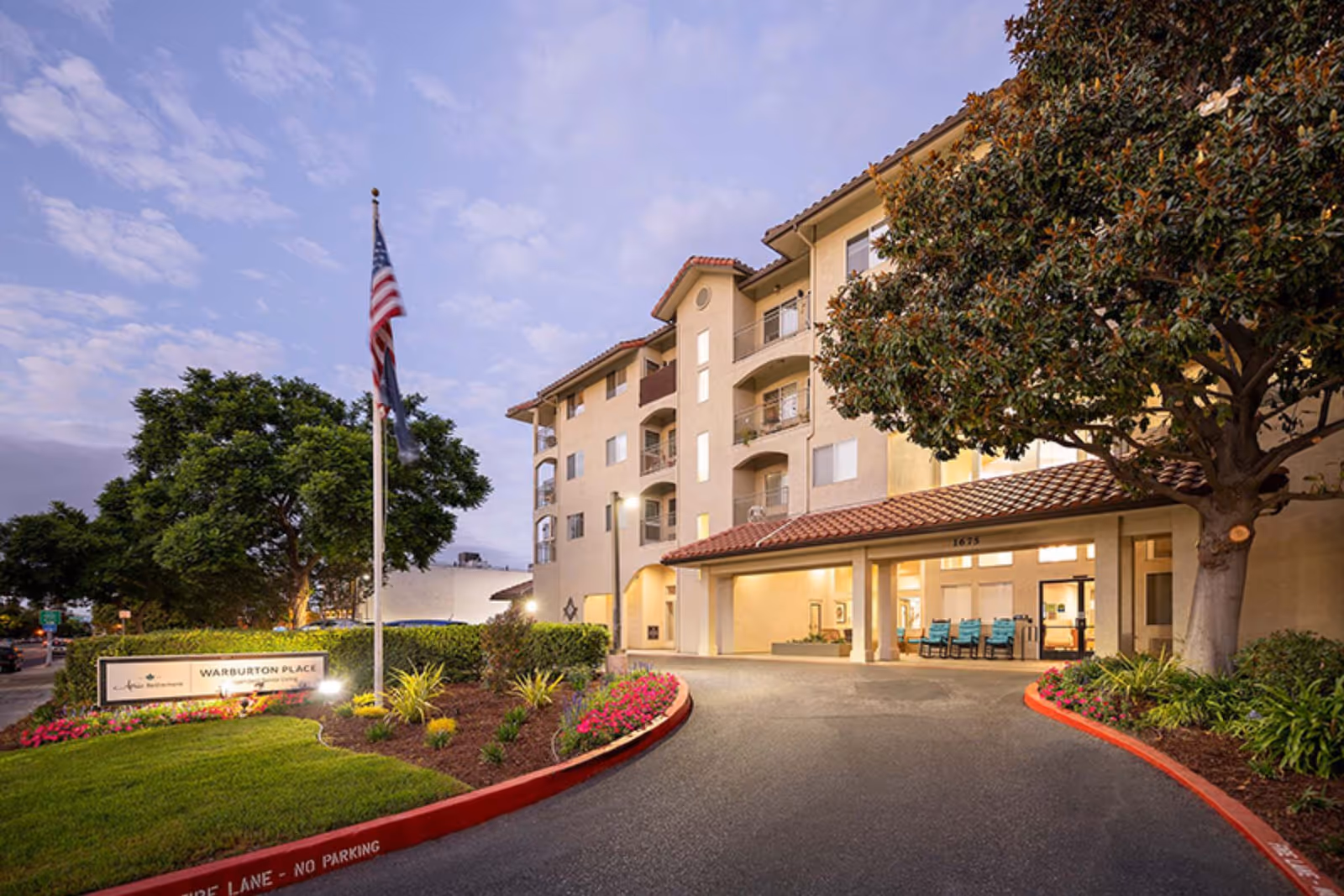 Exterior view of Holiday Warburton Place senior living facility at dusk, showing a multi-story building with balconies, a covered entrance with seating, landscaped greenery, and an American flag on a flagpole near the entrance sign.