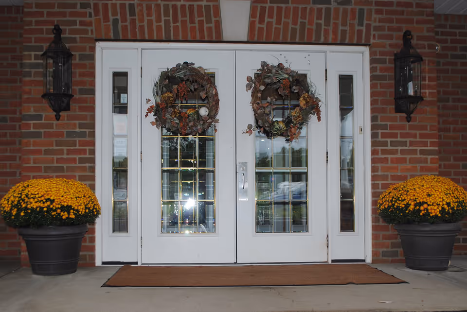 White double entrance doors with glass panes and wreaths set in a brick facade, flanked by lanterns and potted yellow mums on a doormat.
