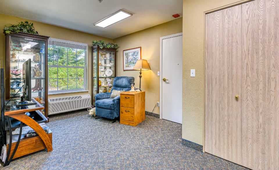 A cozy interior seating area with a blue recliner, wooden side table and lamp, glass display cabinets, TV stand, and a window overlooking greenery.