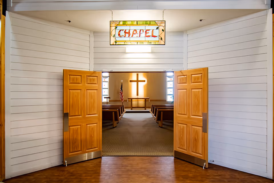 Open wooden double doors leading into a small chapel with pews and a cross at the front beneath a stained-glass sign reading 'Chapel'.