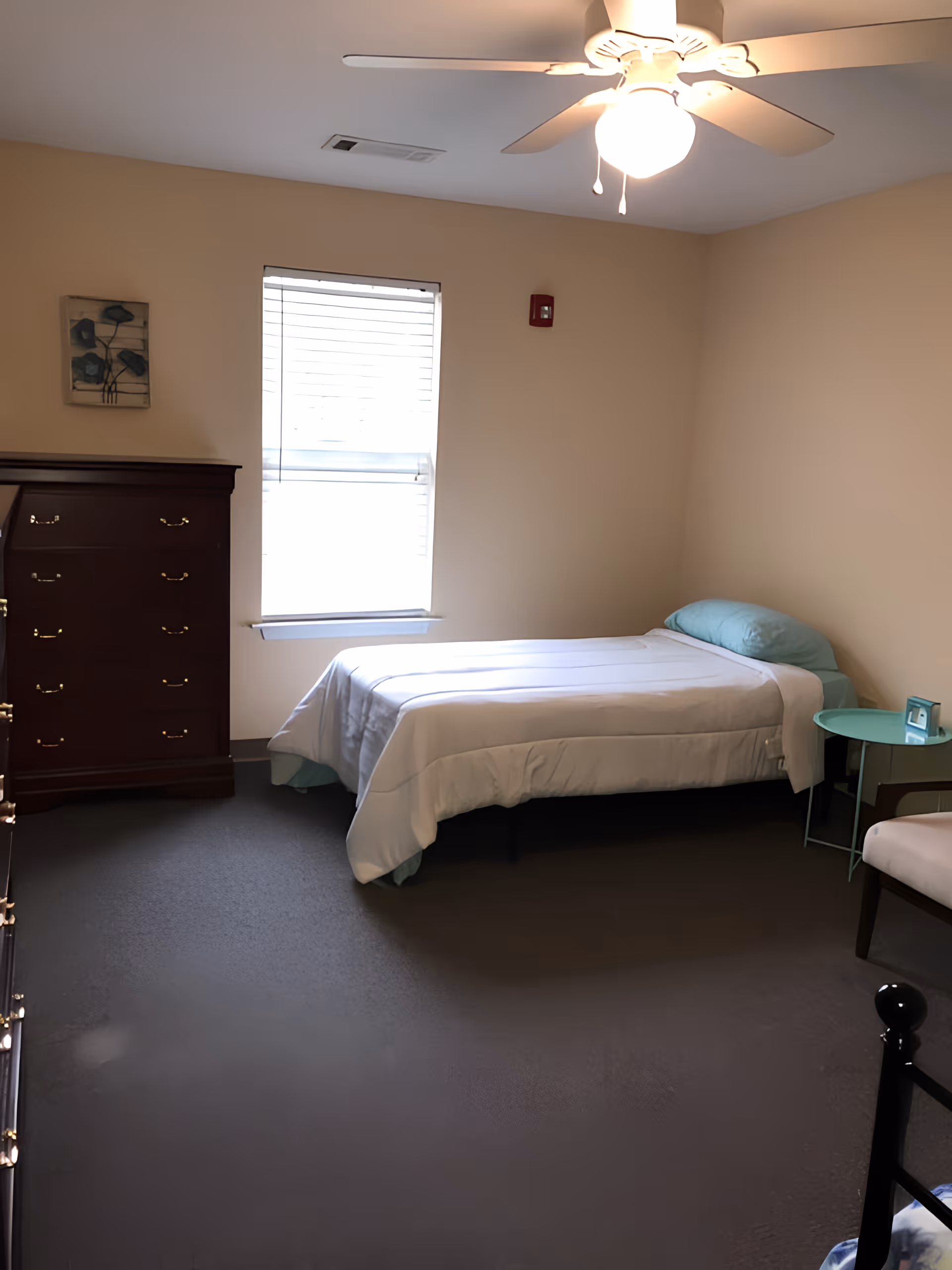 A simple bedroom with a single bed covered in white bedding and a light blue pillow. There is a wooden dresser with multiple drawers on the left side of the room, a small round teal side table next to the bed with a small clock on it, and a beige chair partially visible on the right. The room has beige walls, a window with blinds, a ceiling fan with a light, and dark carpeted flooring.