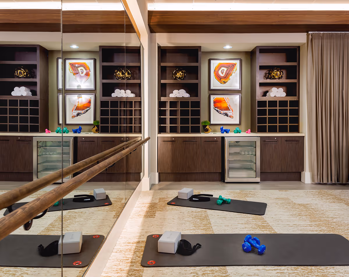 A fitness or exercise room with two black exercise mats on the floor, each equipped with dumbbells, yoga blocks, and straps. The room features a large wall mirror with wooden ballet barre, built-in wooden shelves with rolled white towels, decorative items, and framed abstract artwork. There is also a small refrigerator under the counter. Beige curtains cover a window on the right side.