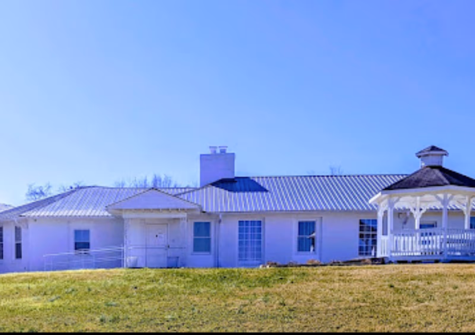 Exterior view of a single-story white building with a metal roof and a white gazebo on the right side, set against a clear blue sky and a grassy lawn in front.