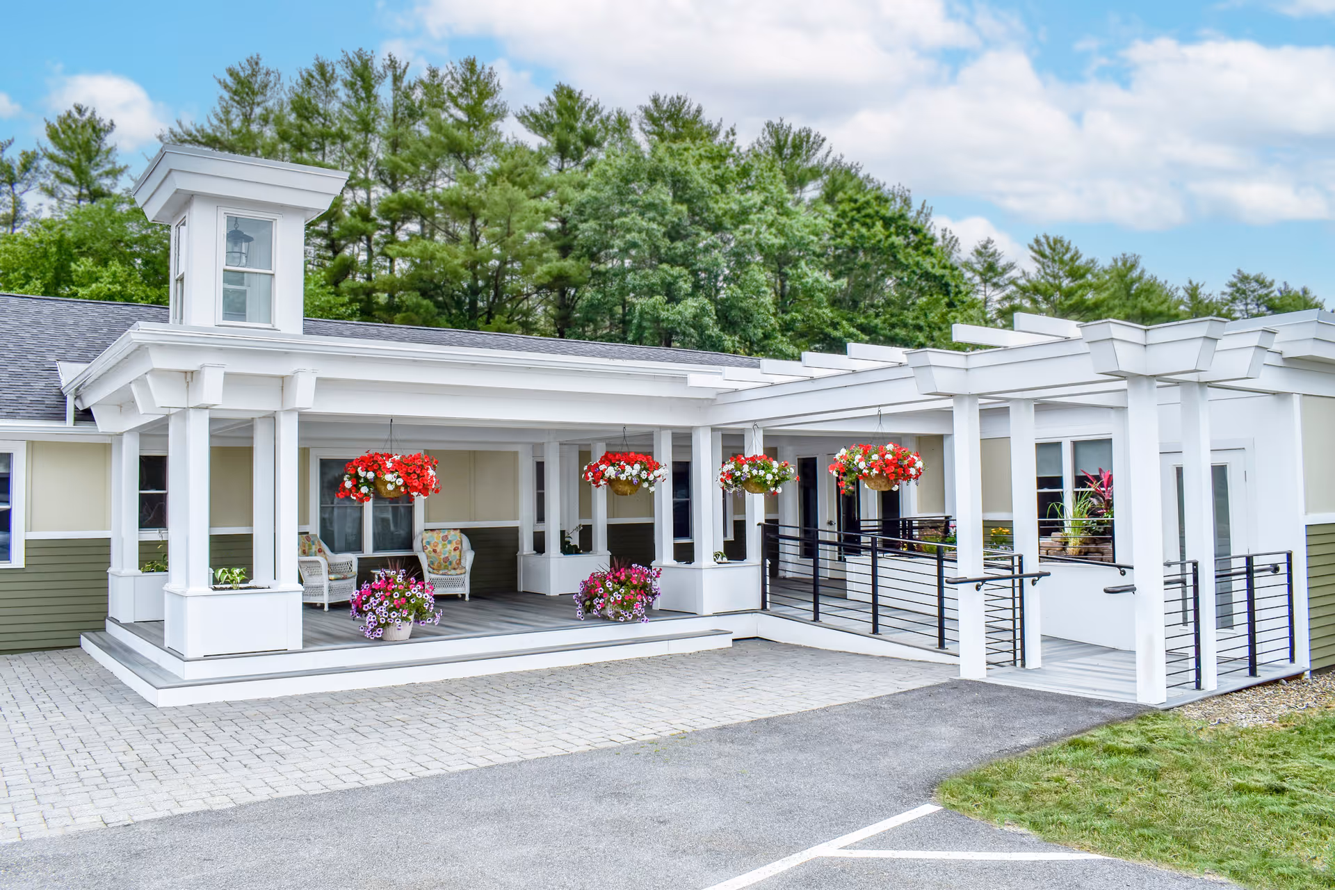 Front entrance of a white memory-care building featuring a covered porch with hanging flower baskets, seating, and an accessible ramp.