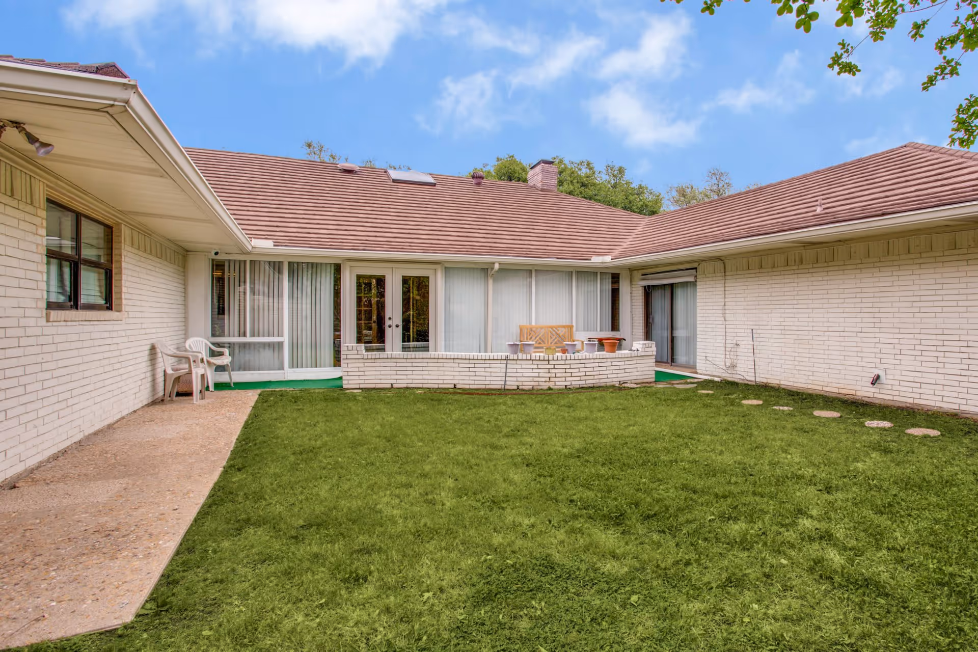 Enclosed grassy courtyard with patio chairs, potted plants and sliding glass doors of a single-story light-brick building under a blue sky.