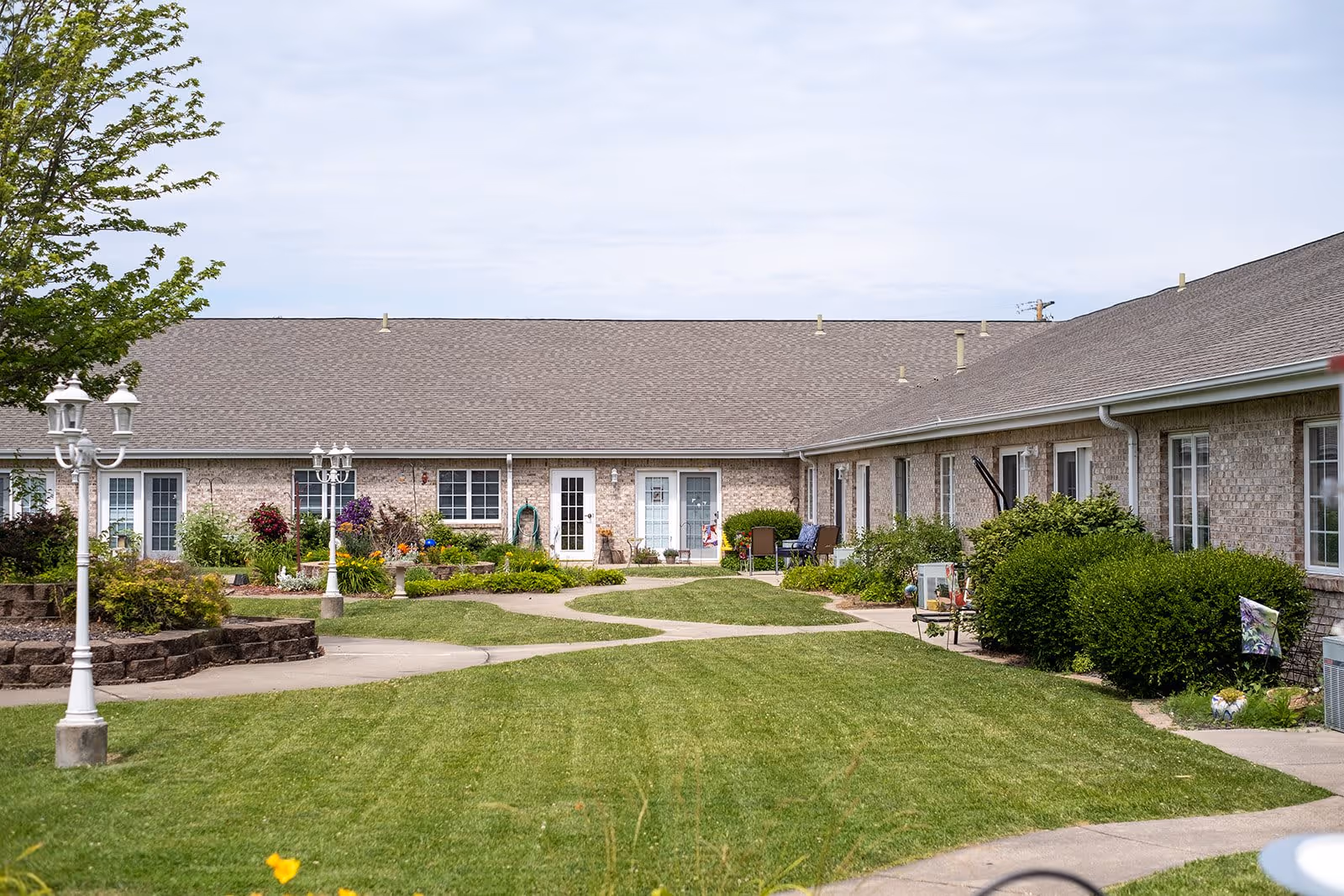 A courtyard area of a senior living facility with a well-maintained lawn, paved walkways, garden beds with flowers and shrubs, and a single-story brick building surrounding the courtyard. There are white lampposts and outdoor seating visible near the building entrances.