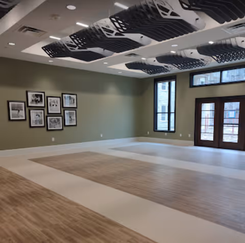 A spacious, empty room with light wood and white flooring, olive green walls, and several black and white framed pictures hanging on one wall. The ceiling features modern black and white acoustic panels and recessed lighting. Large windows and glass doors allow natural light to enter the room.