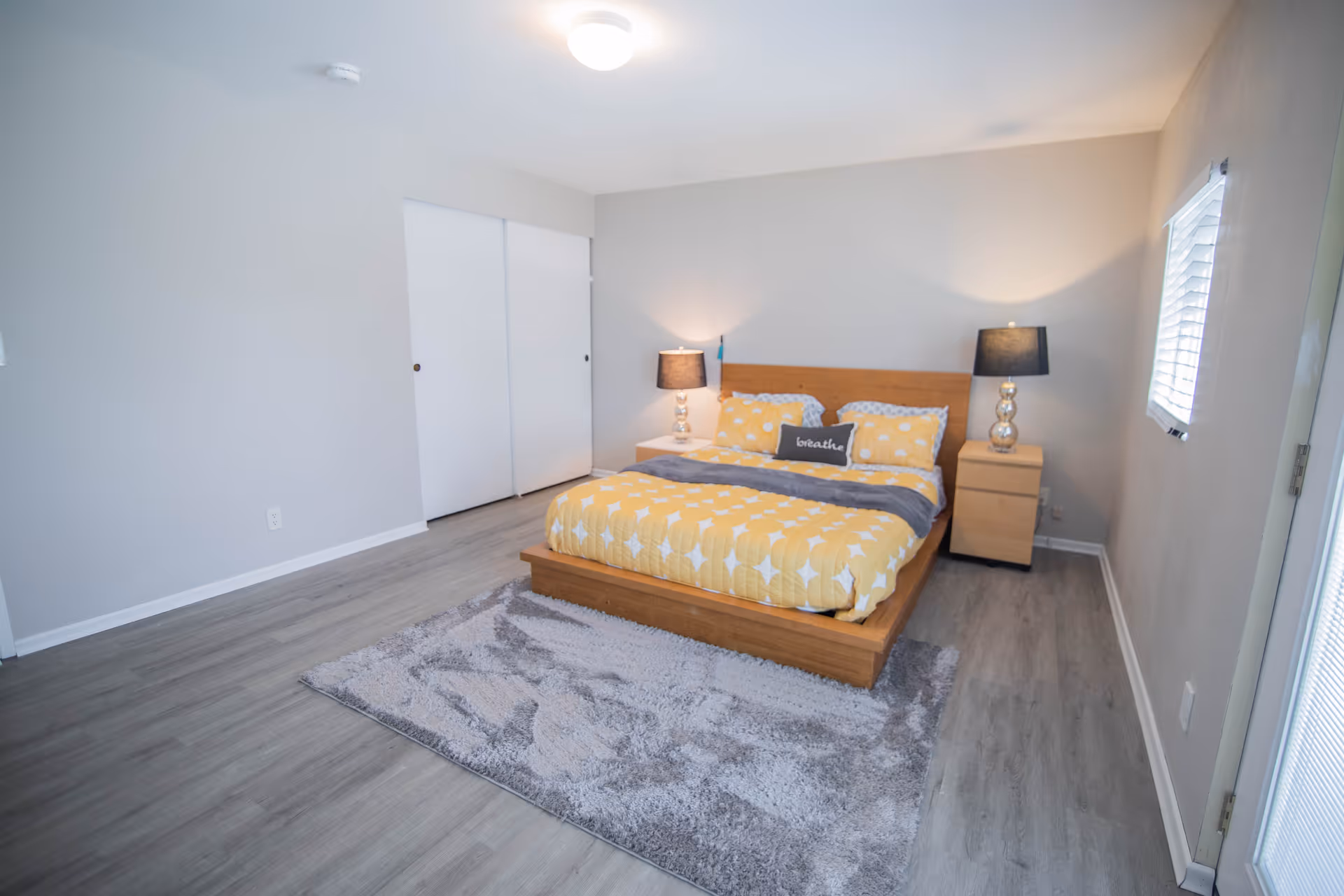 Spacious bedroom with a wooden platform bed dressed in yellow bedding, two bedside tables with lamps, a gray area rug, and a closet with sliding doors.