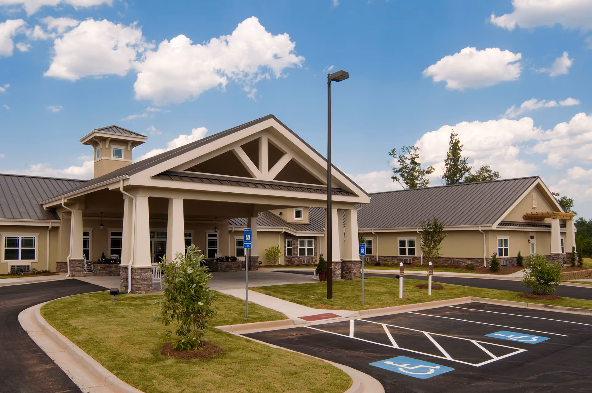 Exterior view of Benton House of Covington, showing a single-story building with a covered entrance supported by columns, a metal roof, and a parking lot with handicap spaces in front. The sky is partly cloudy.