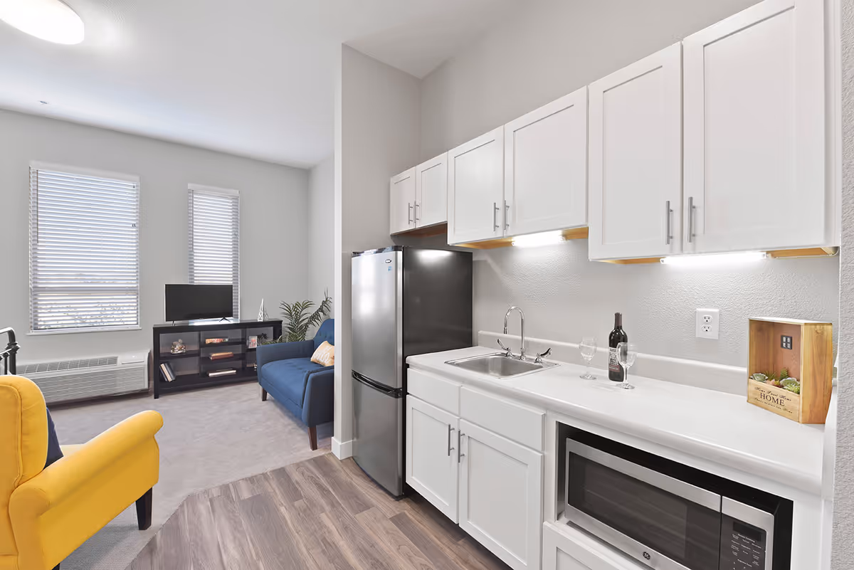 Small open-plan apartment kitchen and living area with white cabinets, a stainless refrigerator, sink and microwave, plus a blue sofa and yellow armchair facing a TV.