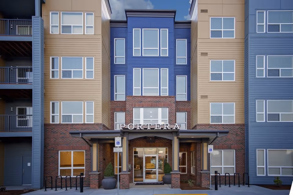 Front exterior view of a multi-story residential building with a central entrance labeled 'PORTERA'. The building features a combination of brick and siding in blue and tan colors, with multiple windows and balconies on the sides. There are handicap parking signs and bike racks near the entrance.