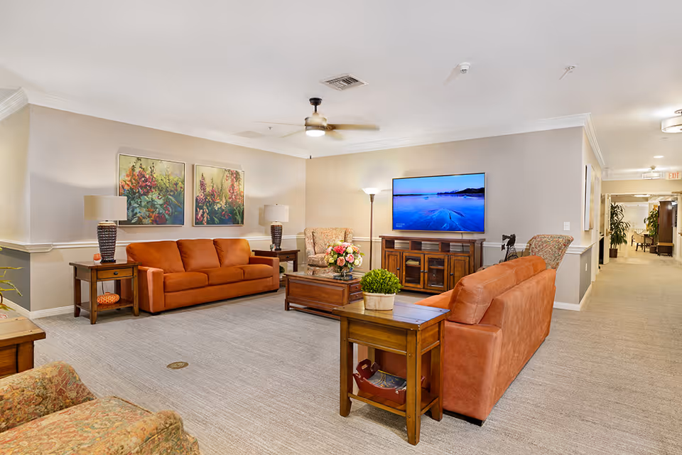 A cozy living room area with two orange sofas, two patterned armchairs, wooden side tables with lamps, a wooden coffee table with a flower arrangement, and a flat-screen TV mounted on the wall. The room has light-colored walls, carpeted flooring, and ceiling fans. There is a hallway visible to the right with plants and additional lighting.