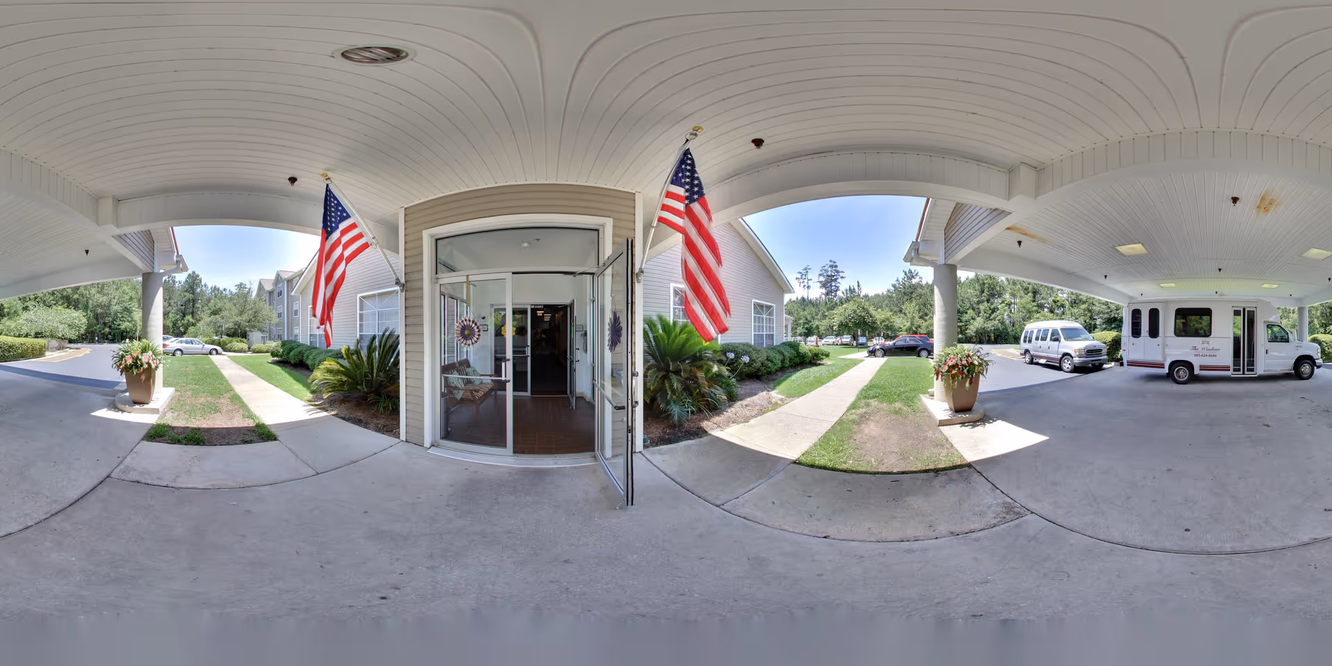 Covered porte-cochere and main entrance with glass doors, American flags, potted plants, landscaping and a parked shuttle van.
