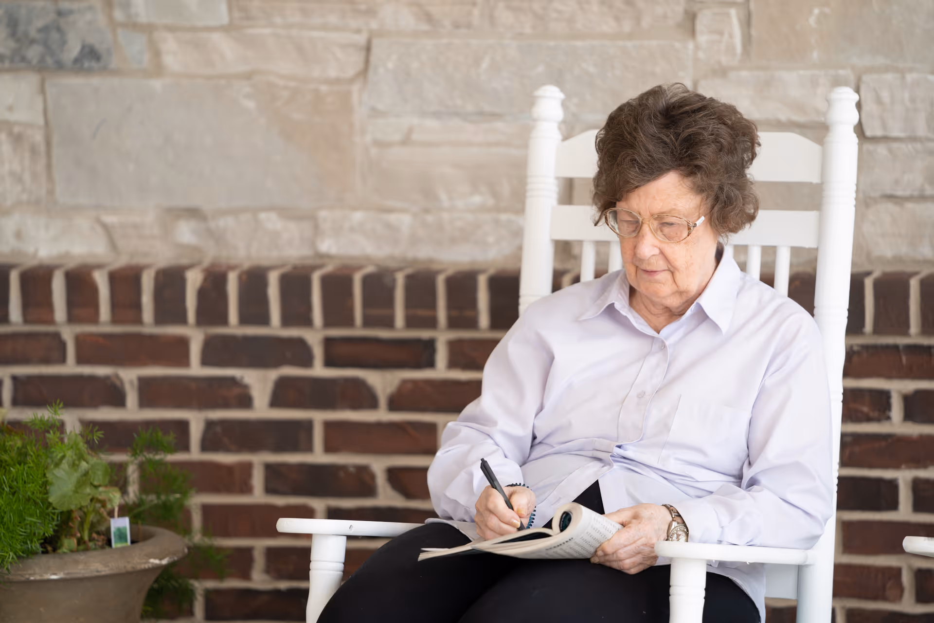 An elderly woman with glasses sitting in a white rocking chair, writing in a notebook or book. She is wearing a light purple shirt and black pants. Behind her is a brick and stone wall, and a potted plant is visible to the left.