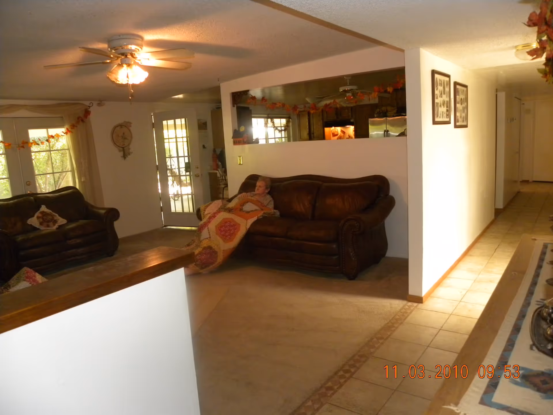 A cozy living room area with two brown leather sofas, one of which has an elderly person sitting and covered with a colorful quilt. The room has a ceiling fan with lights, a tiled floor leading to a hallway, and a large window with curtains decorated with a garland of autumn leaves. The kitchen is partially visible through a large pass-through window in the wall.