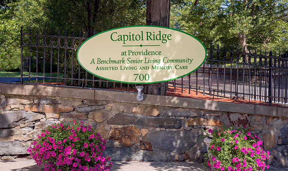 Outdoor sign for Capitol Ridge at Providence, a Benchmark Senior Living Community offering assisted living and memory care, mounted on a stone wall with a black metal fence and surrounded by greenery and purple flowers.