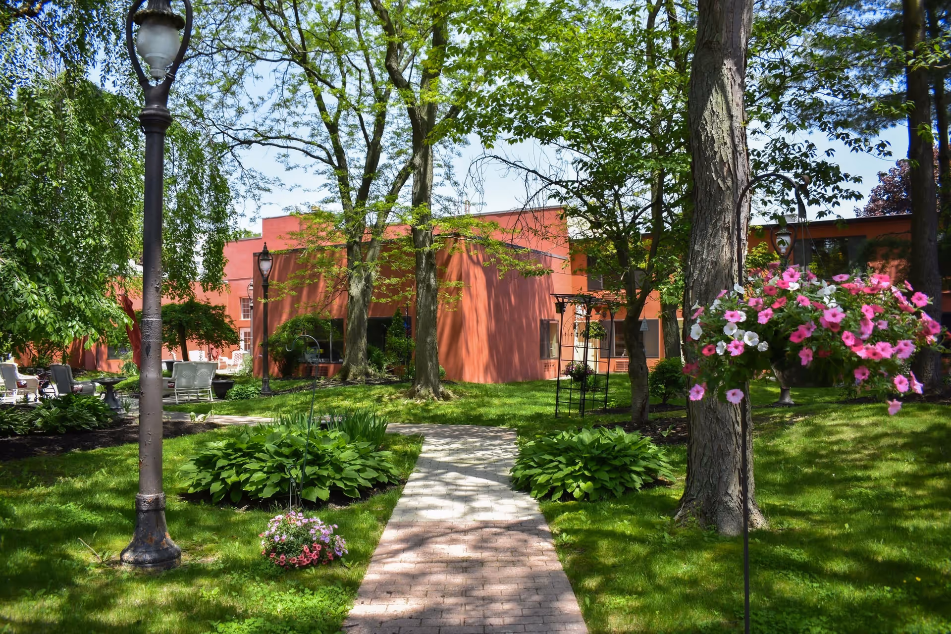 A sunny outdoor garden area with a paved walkway leading towards a red brick building. The garden features green grass, trees, hanging flower baskets with pink and white flowers, and vintage-style street lamps. There are also some outdoor chairs and tables visible to the left side of the image.