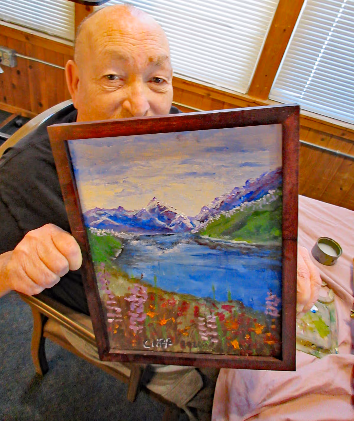 An elderly man sitting indoors holding up a framed painting of a scenic landscape featuring a lake, mountains, and colorful flowers in the foreground.