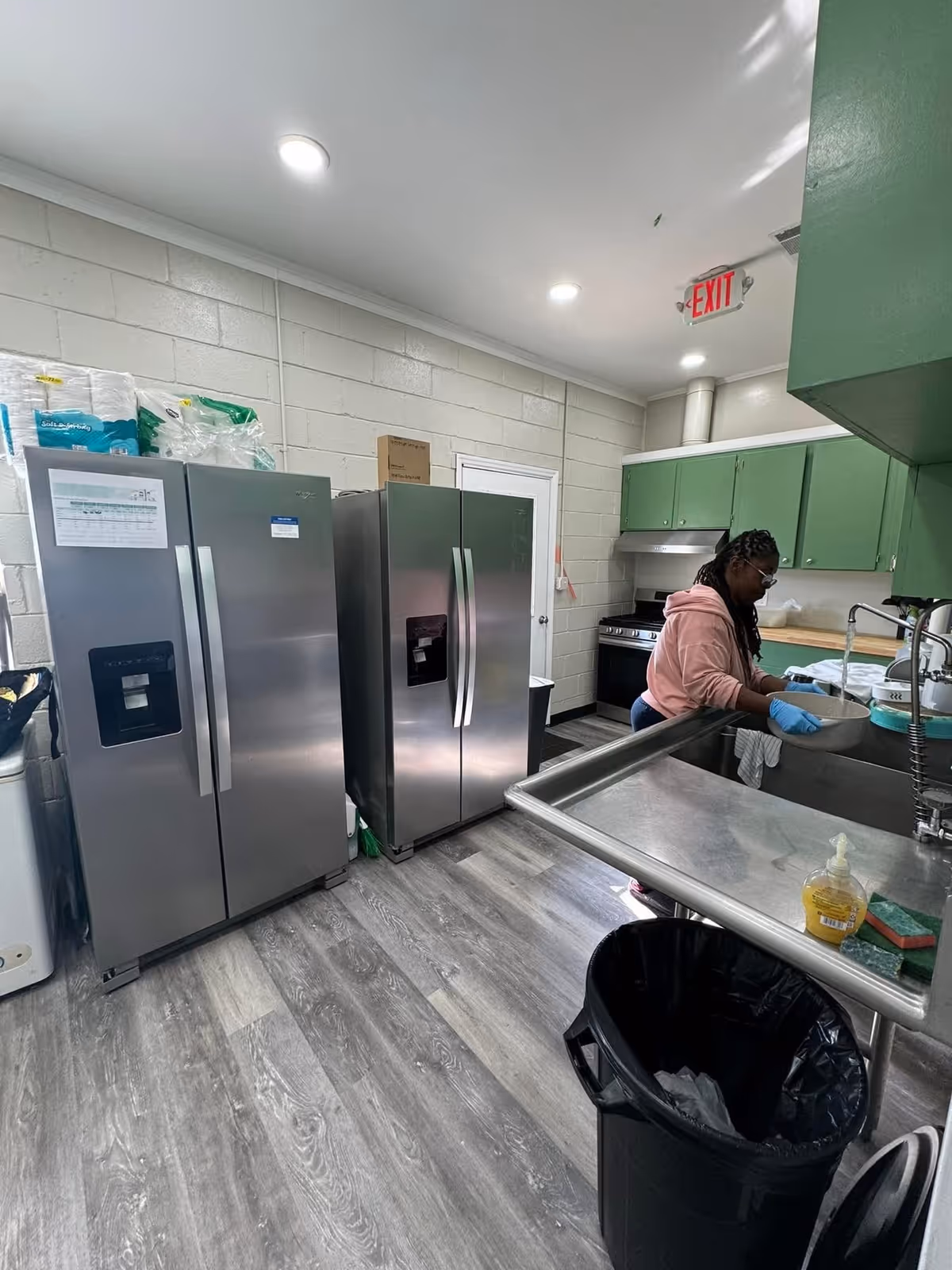 A staff member washes dishes in a kitchen with stainless steel refrigerators, green cabinets, and a large stainless sink.