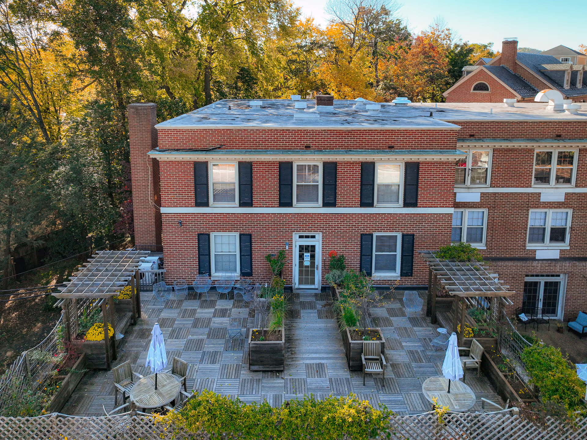 Red brick building with a large wooden terrace patio featuring pergolas, planters, tables, umbrellas, and outdoor seating against a backdrop of trees.