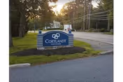Blue entrance sign reading "Cortlandt Healthcare" on a landscaped roadside with trees and power lines in the background.