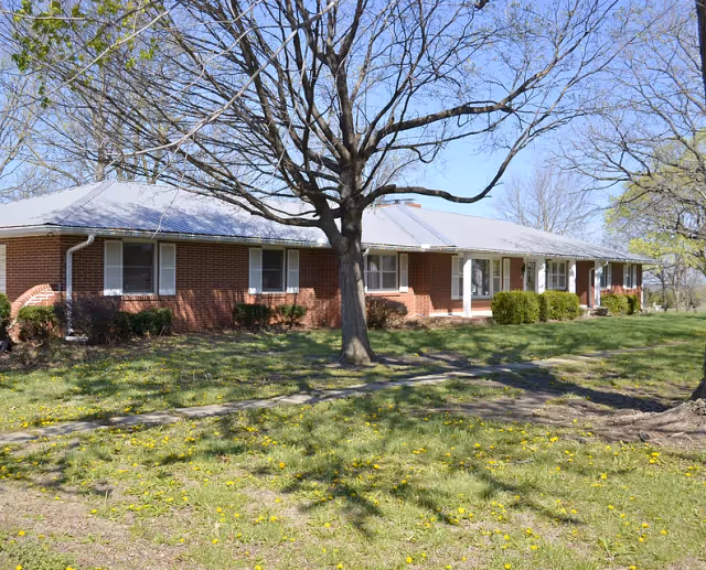 A single-story brick building with a gray metal roof, surrounded by a grassy yard with scattered yellow flowers and leafless trees under a clear blue sky.
