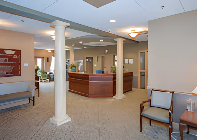 Interior view of a senior living facility reception area with a wooden front desk, beige walls, carpeted floor, two white columns, and seating including a bench and chairs. There are plants and framed items on the walls.