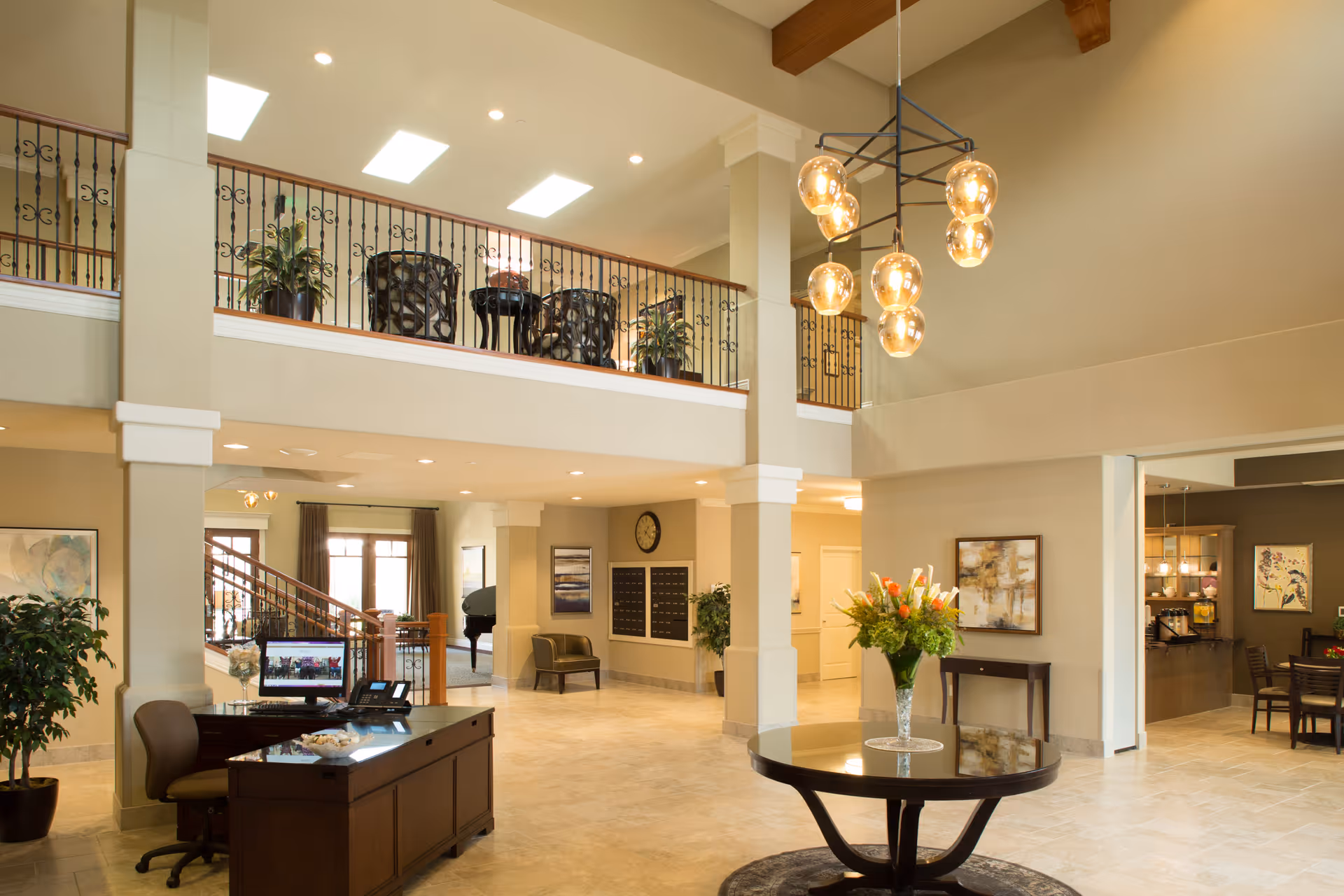 Spacious and well-lit interior lobby area of a senior living facility with a reception desk, a round table with a flower arrangement, seating areas, and a staircase leading to an upper balcony with additional seating and plants.