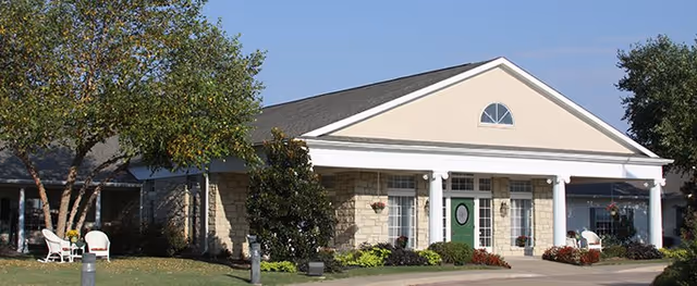 Single-story assisted living facility building with a beige exterior, white columns at the entrance, a green door, and surrounding trees and shrubs under a clear blue sky.