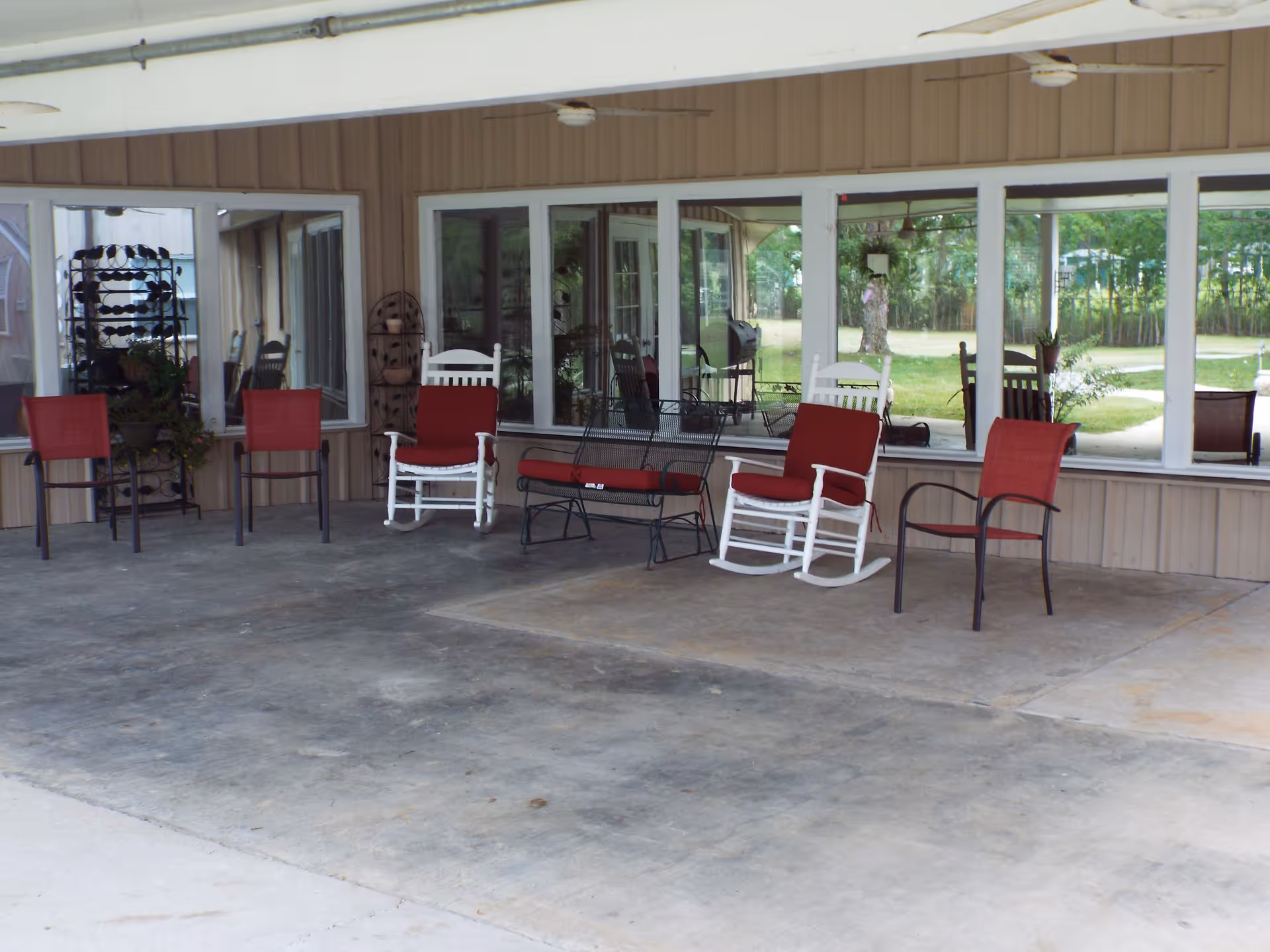 Covered outdoor patio area with several chairs including red cushioned metal chairs, white wooden rocking chairs with red cushions, and a black metal bench with a red cushion. The patio is adjacent to a building with large windows reflecting greenery and outdoor scenery.