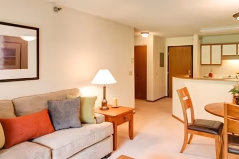 A cozy living room area with a beige sofa adorned with colorful cushions, a wooden side table with a lamp, and a framed picture on the wall. Adjacent to the living room is a small dining area with a round wooden table and chairs, and a kitchen with wooden cabinets and a countertop.