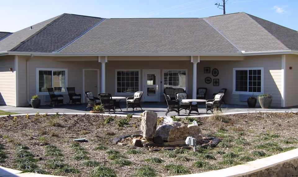 Front patio of a single-story care facility with wicker chairs, tables, and a small landscaped rock garden.