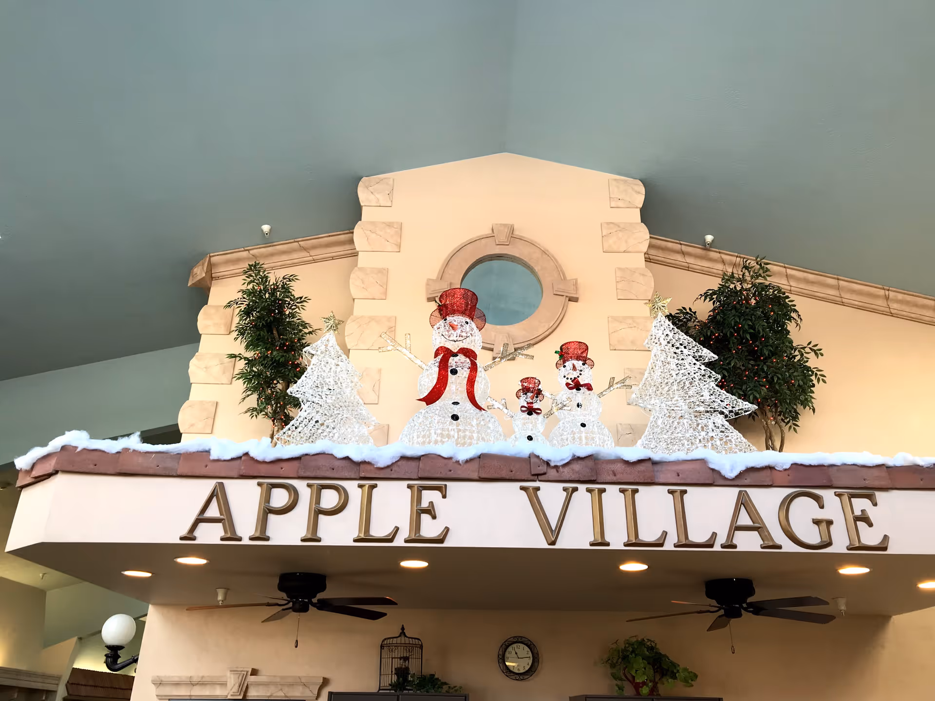 Interior view of a building area with a decorative ledge displaying lighted snowmen and Christmas trees, with the words 'APPLE VILLAGE' in large gold letters below. The ceiling is painted blue, and there are ceiling fans and plants visible underneath.
