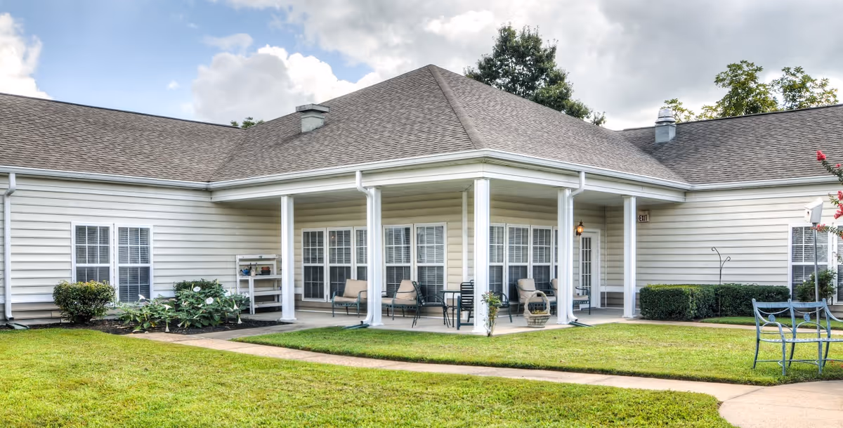 Exterior view of a single-story senior living facility building with beige siding and a gray shingled roof. The building features a covered patio area with several chairs and small tables. There are well-maintained green lawns and shrubs surrounding the building under a partly cloudy sky.