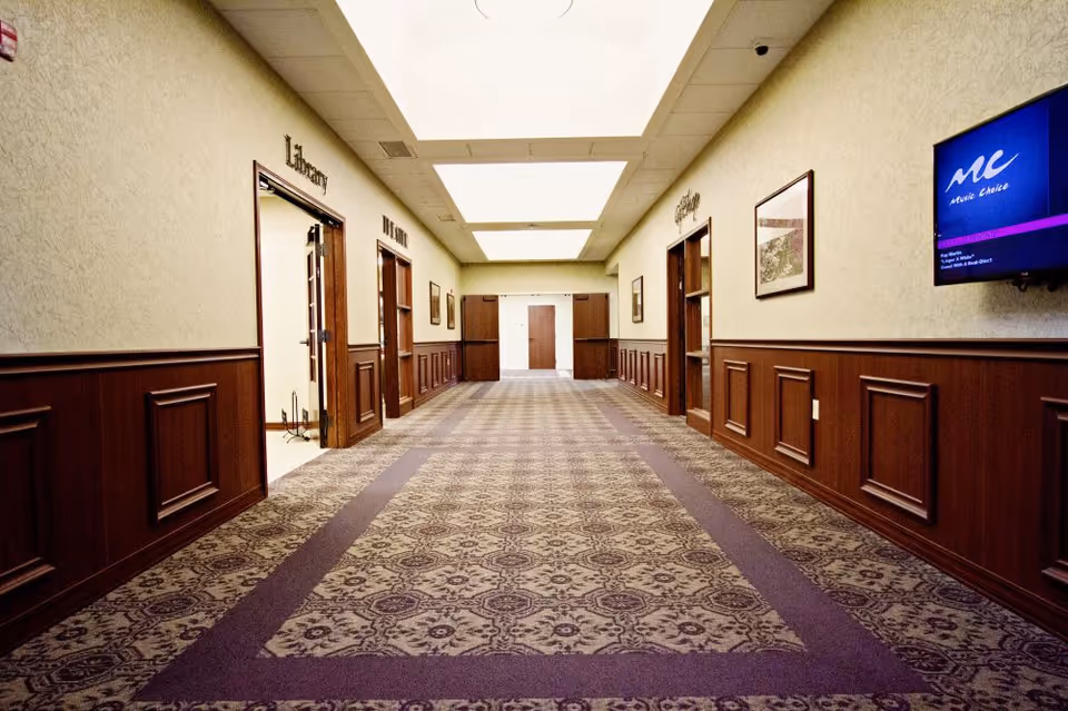 A well-lit hallway with patterned carpet and wood-paneled lower walls. Doors on the left and right lead to rooms labeled Library, The Loft, and Coffee. A television screen is mounted on the right wall displaying a music channel.