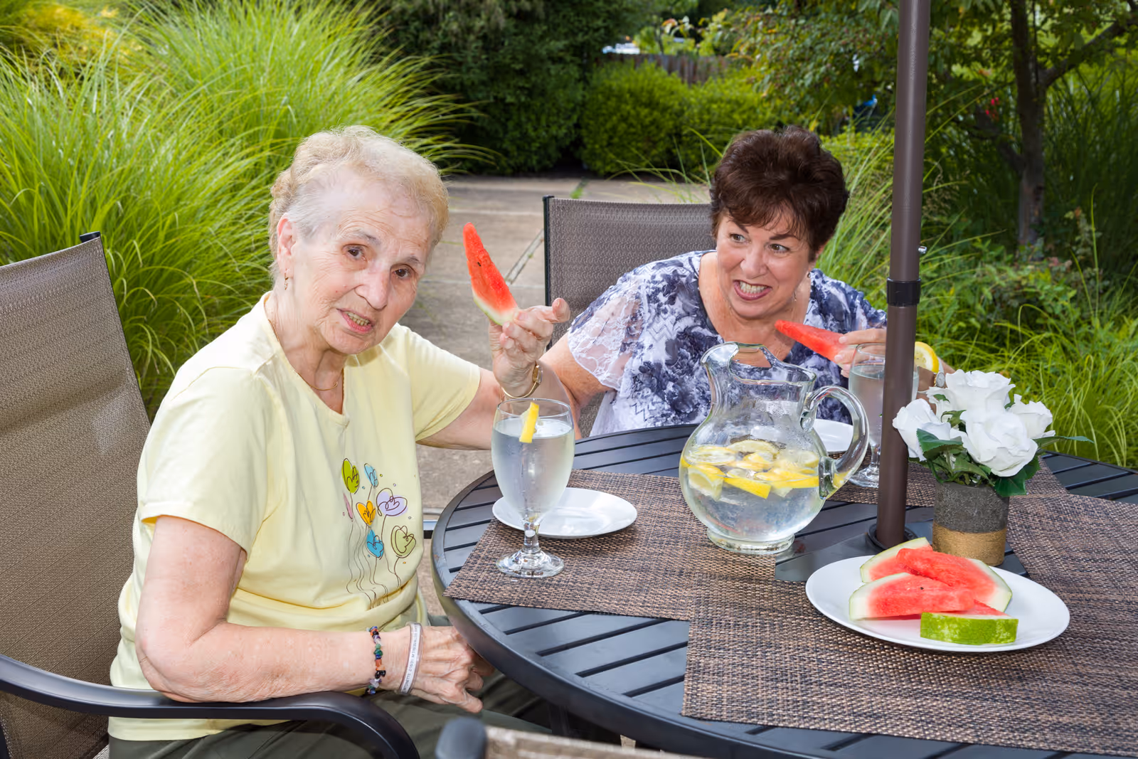 Two elderly women sitting at a round outdoor table with a pitcher of lemon water, glasses of water with lemon, and a plate of watermelon slices. They are enjoying watermelon slices and surrounded by green plants and bushes.