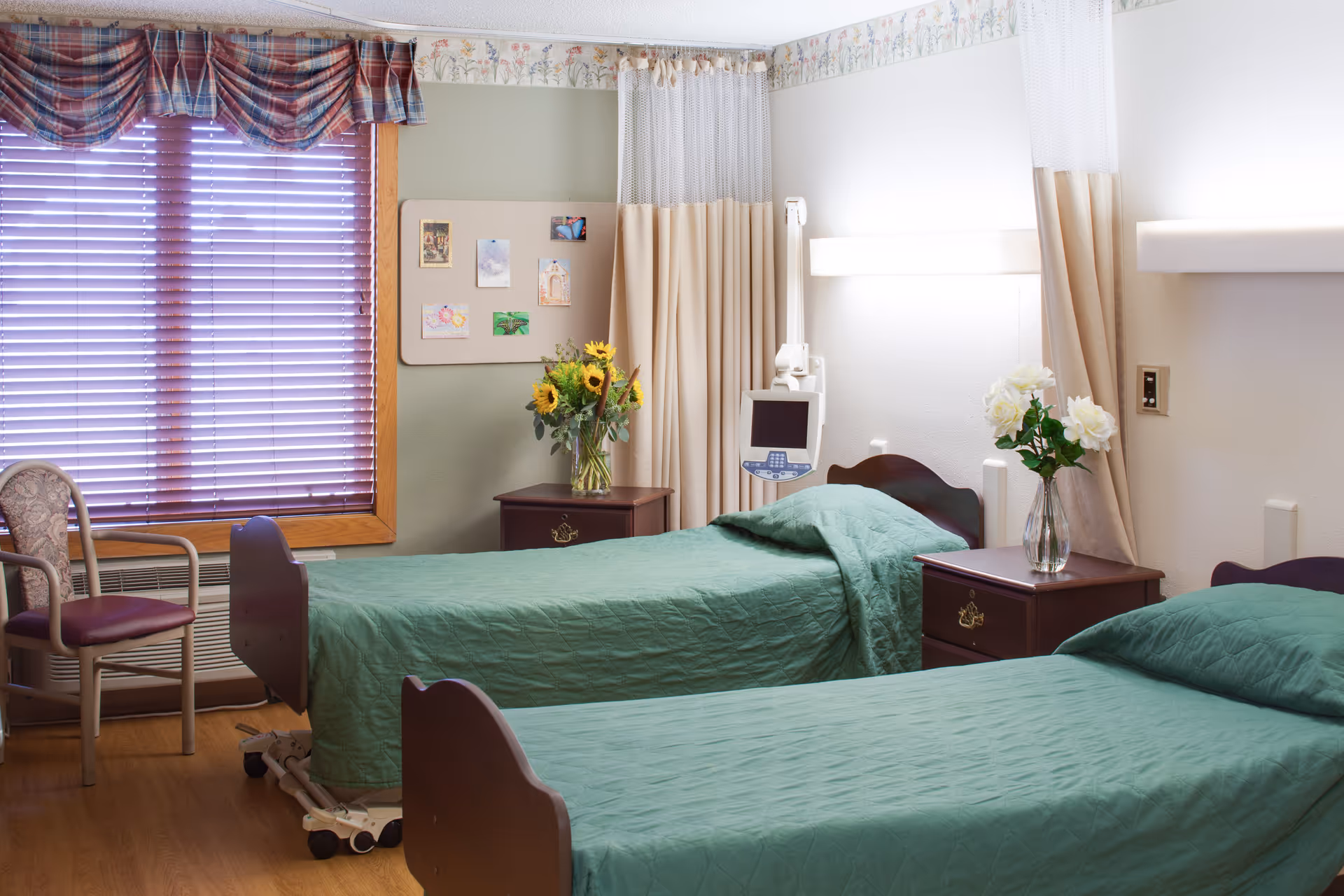 Two twin beds with green bedspreads in a bright assisted-living bedroom with nightstands, flowers, and a window with blinds.
